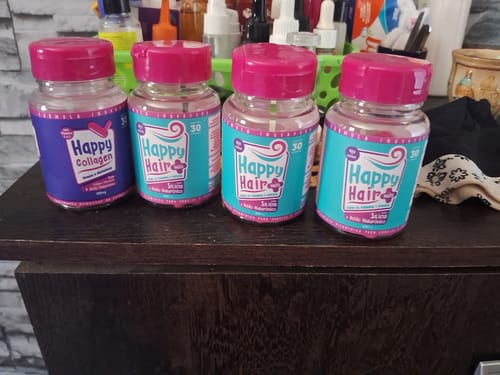 Cliente exibe os três potes de Happy Hair e um pote de Happy Collagen recebidos no kit, alinhados sobre uma mesa de madeira.