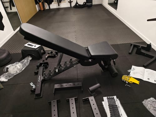 Ein Rezensent baut die schwarze B6 Hantelbank in einem Fitnessraum auf; Teile und Anleitungen liegen auf dem Boden.