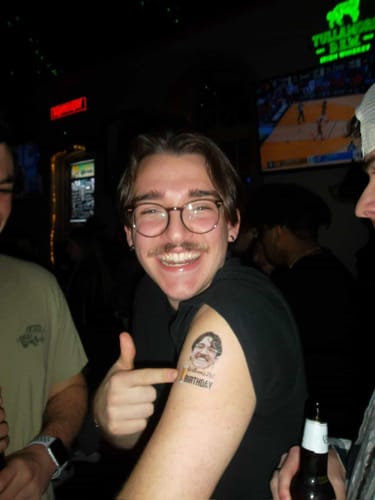 Customer smiling and pointing to a custom birthday tattoo on their bicep at a bar.