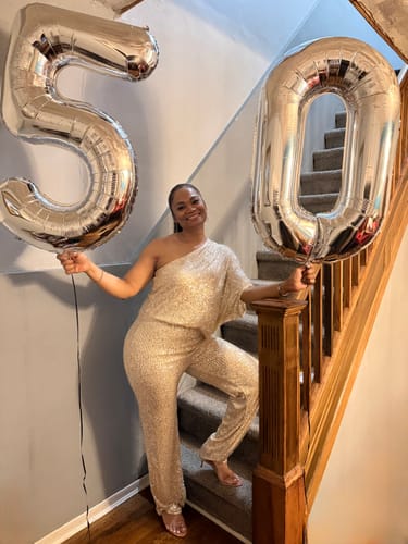 Reviewer smiling on a staircase in the Rebecca Champagne Gold One Sleeve Sequin Jumpsuit, holding large '50' balloons.