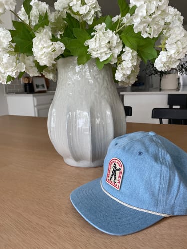 Customer's light blue Big Flat Bill Unstructured Rope Hat resting on a wooden table next to a vase.