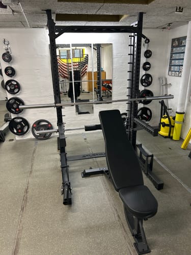 Rezensent zeigt das schwarze R7 Saber Squat Rack, das mit einer Hantelbank in einem Heim-Fitnessstudio aufgestellt ist.