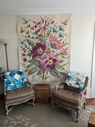 Reviewer's finished Tilda Flower Branch quilt hanging on a wall above two chairs, showing the colorful, pixelated floral design.