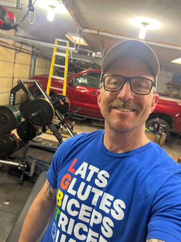 Customer wearing the blue Muscle Alphabet T-Shirt while taking a selfie in a home gym.