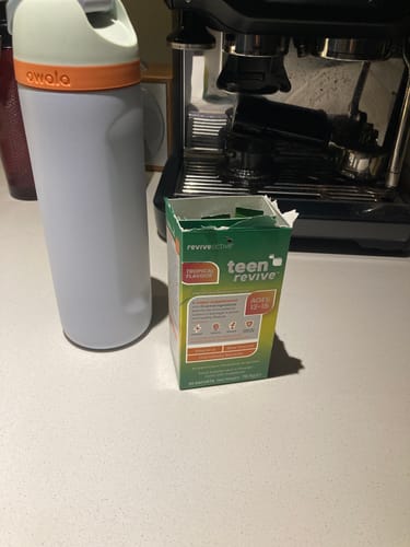 Reviewer's open box of Teen Revive Tropical Flavour sachets on a kitchen counter next to a water bottle.