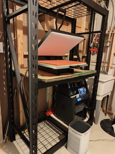 Customer's Bambu Lab P1S Combo 3D printer sitting on a black steel shelf below a shirt press in a workshop.