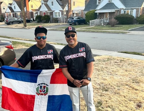 Two customers wearing matching black 'Dominicano' baseball jerseys while holding a large Dominican Republic flag outdoors.
