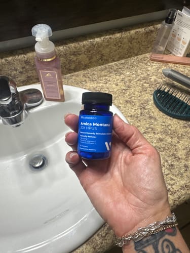 Customer holding the blue bottle of Arnica Montana 30X HPUS tablets in their bathroom.