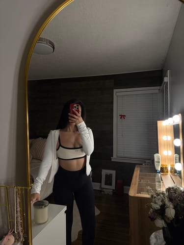 Reviewer taking a mirror selfie wearing the cream and black Micro Bra with black leggings and an open white cardigan.