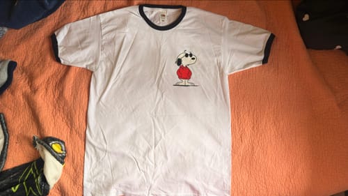 Customer's white ringer t-shirt with black trim and a Snoopy graphic on the chest, laid flat on an orange surface.