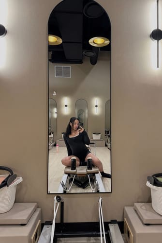 Reviewer taking a mirror selfie on a Pilates reformer while wearing a black top with cream trim.