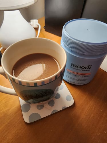Reviewer's prepared mug of Hot Chocolate next to the product's light blue container on a wooden desk.