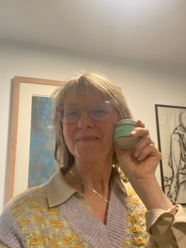 Customer smiling while holding the tin of Glow to Bed cream next to her cheek.