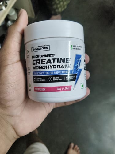 A reviewer holds a white tub of Wellcore Micronised Creatine Monohydrate in the Fruit Fusion flavor.
