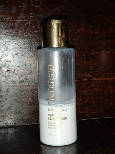 Customer's bottle of Dream Glaze Peptide Milk Toner, with its gold cap, on a dark wooden surface.