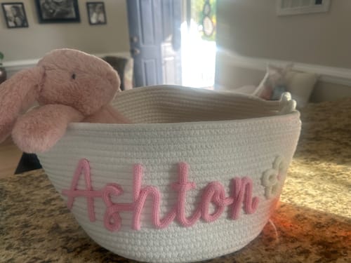 Customer's white knitted basket personalized with 'Ashton' in pink yarn and a flower icon, holding a plush bunny.