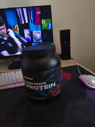 Reviewer's tub of CFM Whey Protein on a desk in front of a computer monitor and keyboard.