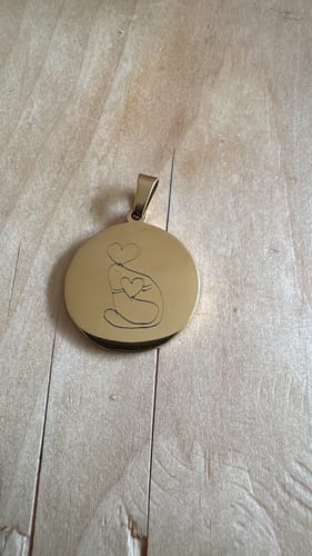 Kundenbewertung: Goldfarbener Anhänger mit der Gravur einer stilisierten Figur mit Herzen auf einer Holzoberfläche.