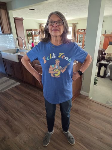 Reviewer standing with hands on hips, showing the fit of the blue Folk Yeah Tee in a home setting.