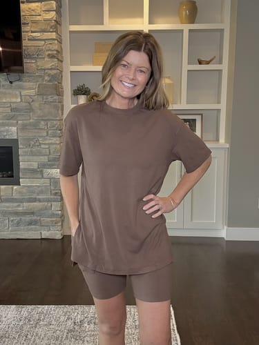 Clienta posando en su sala de estar con el cómodo conjunto LumiSet™ de camiseta oversized y shorts en color marrón.