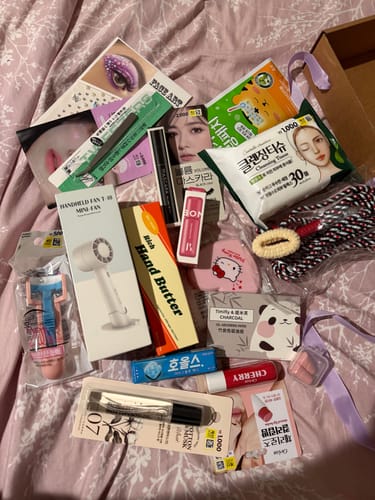 Reviewer displays items from the K-POP CONCERT ESSENTIAL KIT, including a mini fan, mascara, and other cosmetics.