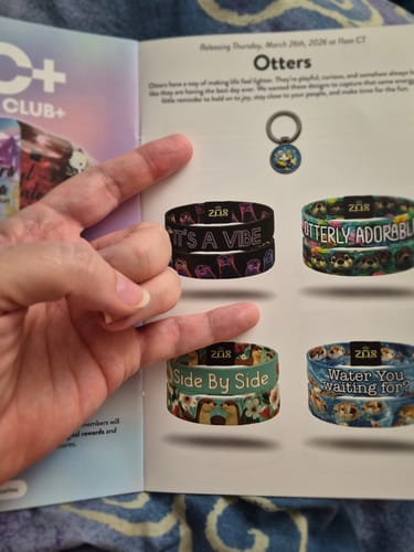 A customer's hand holding the March 2026 Booklet open to a page showing several new otter-themed product designs.
