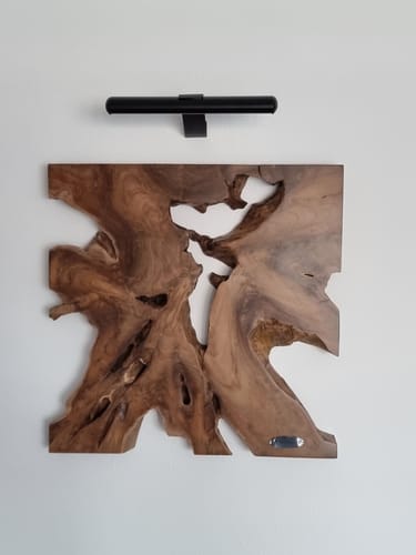 Kunde zeigt das Wandbild NAGA aus Teakholz an einer weißen Wand, mit einer schwarzen Bildleuchte darüber montiert.
