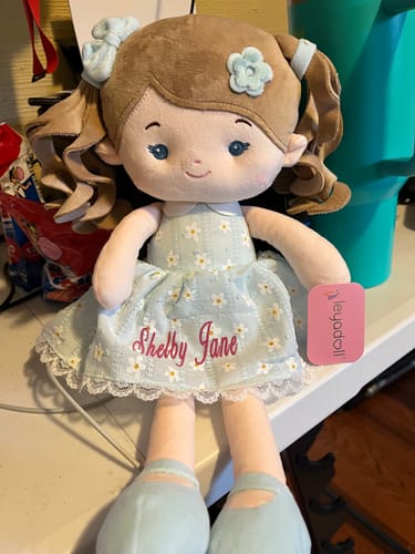 Customer's personalized Leya Doll sitting upright, showing the name 'Shelby Jane' embroidered in pink on its blue daisy-print dress.