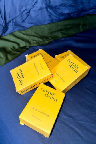 Reviewer displays five yellow boxes of the Turmeric Fade & Glow Bar on a blue surface.