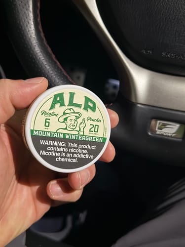 Reviewer holding a tin of ALP Mountain Wintergreen Nicotine Pouches in a car.