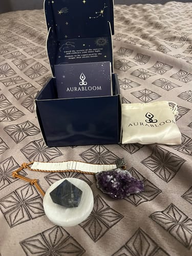 Customer's AuraBloom Subscription Box opened on a bed, showing a selenite dish, a bracelet, and various crystals.