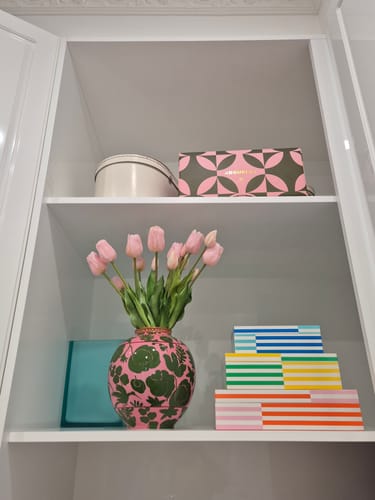 Customer's pale pink tulips in a decorative vase on a shelf inside a white cabinet.