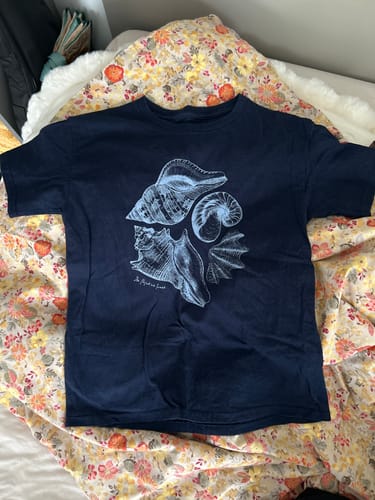 Customer's dark blue sandy baby tee with a seashell graphic print laid flat on a bed.