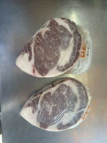 Reseñador muestra dos filetes de Rib Eye Steak Prime crudos y envueltos individualmente en plástico sobre una superficie metálica.