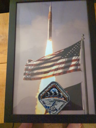 Customer's Artemis II - Collectors Patch in a black frame, mounted over a photo of a rocket launching with a US flag.