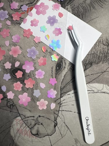 Reviewer holding a tiny floral sticker with the white Sticker Tweezer, with more stickers nearby.