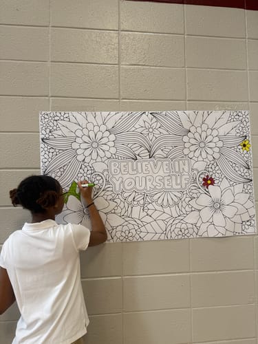 A reviewer coloring the large Zentangle 'Believe In Yourself' sheet which is taped to a cinderblock wall.