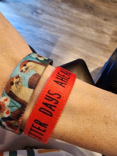 Close-up of a customer's wrist wearing the Side By Side Bracelet with its otter design next to a red bracelet.