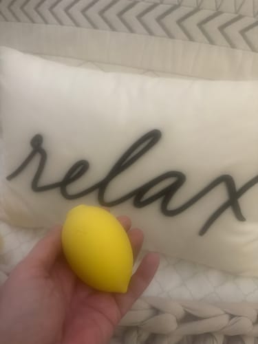 Customer's hand holding the yellow Lem Clitoral Massager in front of a white pillow with 'relax' on it.