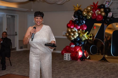 Reviewer smiling in the Rebecca Champagne Gold One Sleeve Sequin Jumpsuit while holding a microphone at an event.