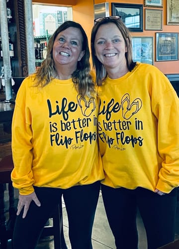 Two reviewers smiling in their matching yellow 'Life Is Better In Flip Flops' graphic crewneck sweatshirts.