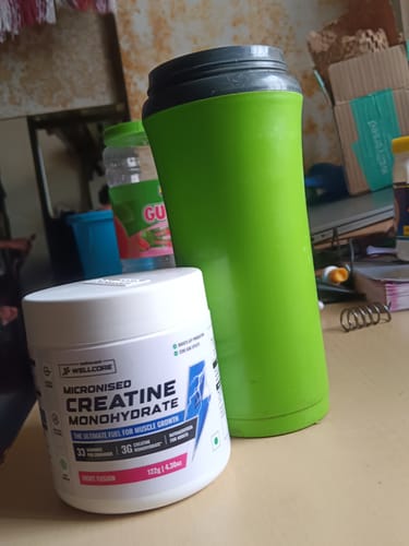 Reviewer's tub of Wellcore Creatine Monohydrate on a desk next to a green shaker bottle.