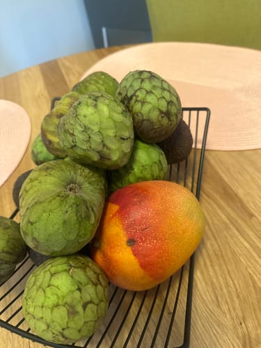 Kundenrezension: Eine große, farbenfrohe Mango aus der Mix-Box liegt in einem schwarzen Korb mit anderen Früchten.