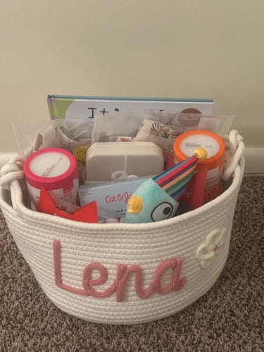 Customer's personalized white knitted basket with the name 'Lena' in pink yarn and a flower icon, filled with gifts.