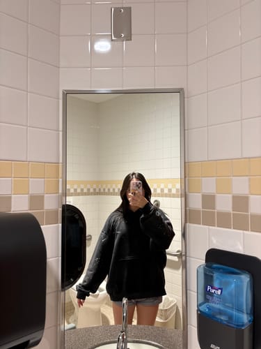Reviewer taking a mirror selfie, showing the oversized fit of the black Arirang hoodie in a restroom.