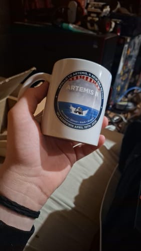 A reviewer's hand holding the white 'Apollo to Artemis' mug, showing the mission patch graphic.