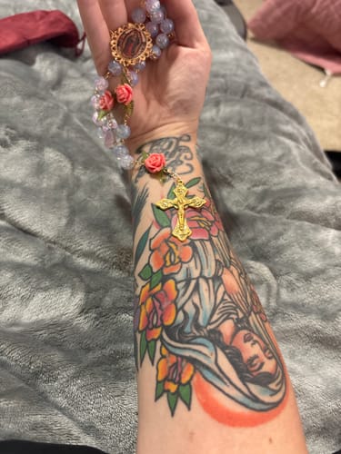 Reviewer displays the Virgin Mary rosary with pastel beads and pink roses on their tattooed arm.