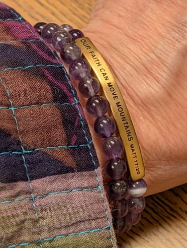Customer's wrist wearing a purple STACKER - AMETHYST beaded bracelet next to a gold-colored engraved band on a patterned sleeve.