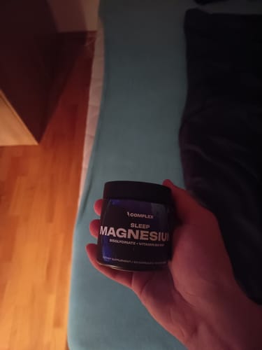 Customer's hand holding a black jar of Sleep Magnesium supplement in a bedroom.