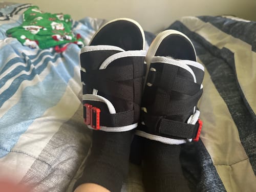 Customer wearing black Mystery DracoSlides with white trim and red buckles over black socks while sitting on a bed.